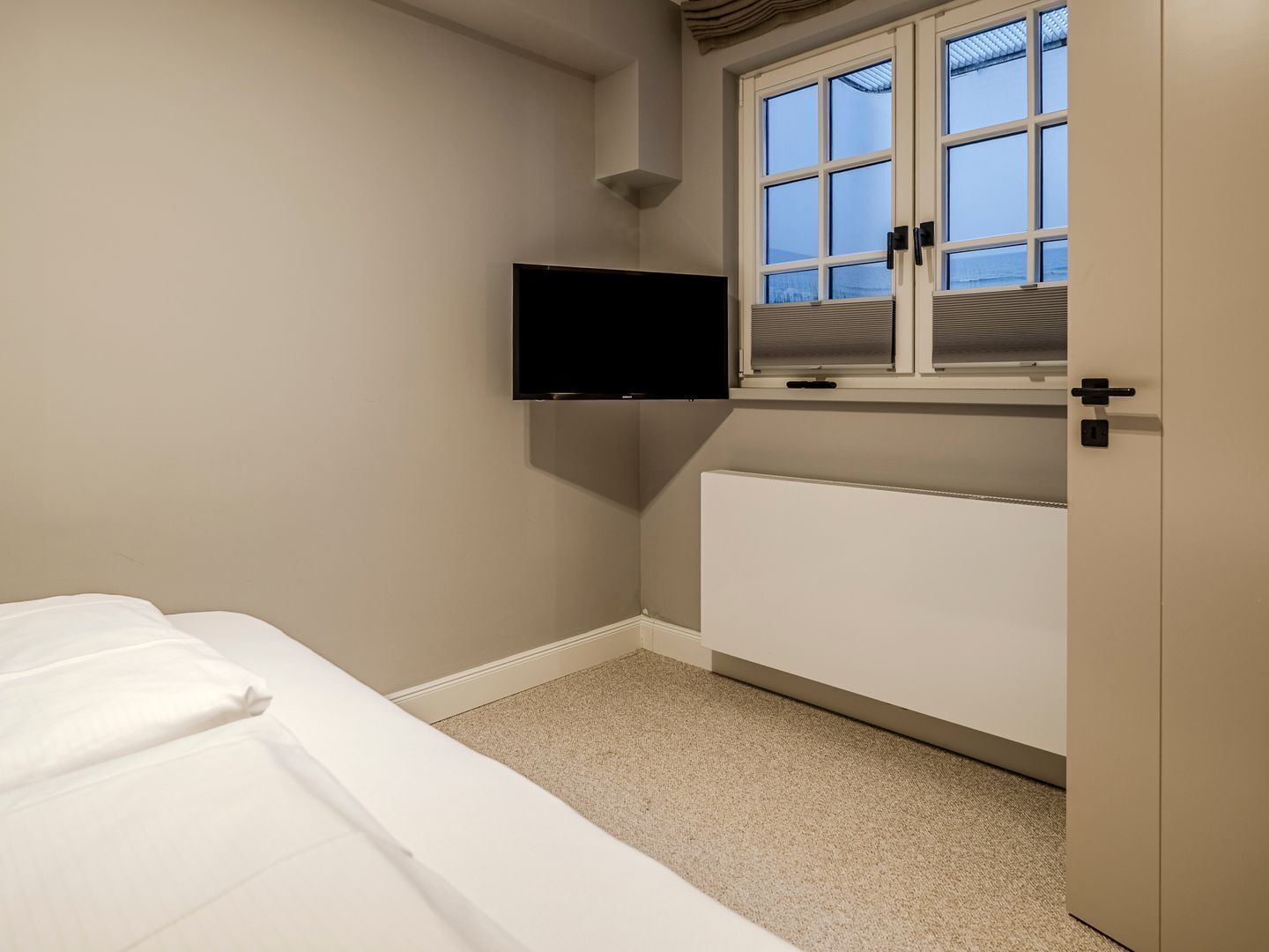 Quarto com cama, televisão na parede, janela com persianas e radiador.