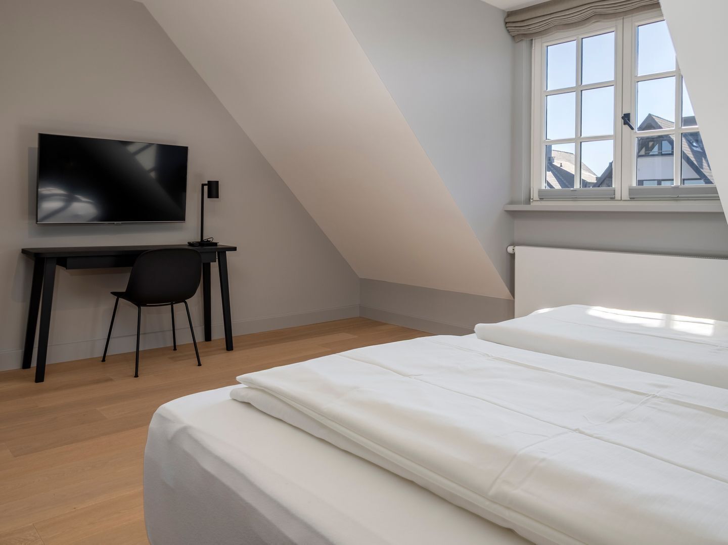 Quarto moderno e minimalista com teto inclinado, uma janela ampla, uma televisão de tela plana e uma escrivaninha com cadeira.