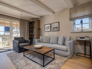 En moderne stue med en grå sofa, en grå lænestol, et træbord og en hvid skål. To billeder hænger på væggen, der er en gulvlampe, og et vindue med udsigt til bygninger.