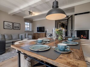 Un coin repas avec une table en bois dressée pour quatre personnes, un grand luminaire suspendu et une cheminée en arrière-plan.