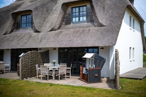 A thatched-roof house with a dining area, a small outdoor seating area, and a large window.