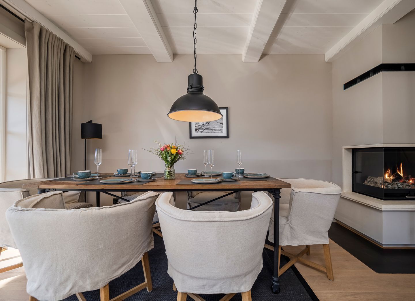 A dining area with a wooden table set for four, surrounded by four chairs. A black pendant light hangs above the table.
