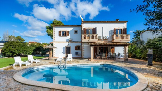 Une maison à deux étages avec une piscine, du mobilier d’extérieur et un ciel d’un bleu éclatant.