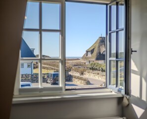 A view through a window of a coastal area with thatched-roof houses, a stone wall, and a parked car.