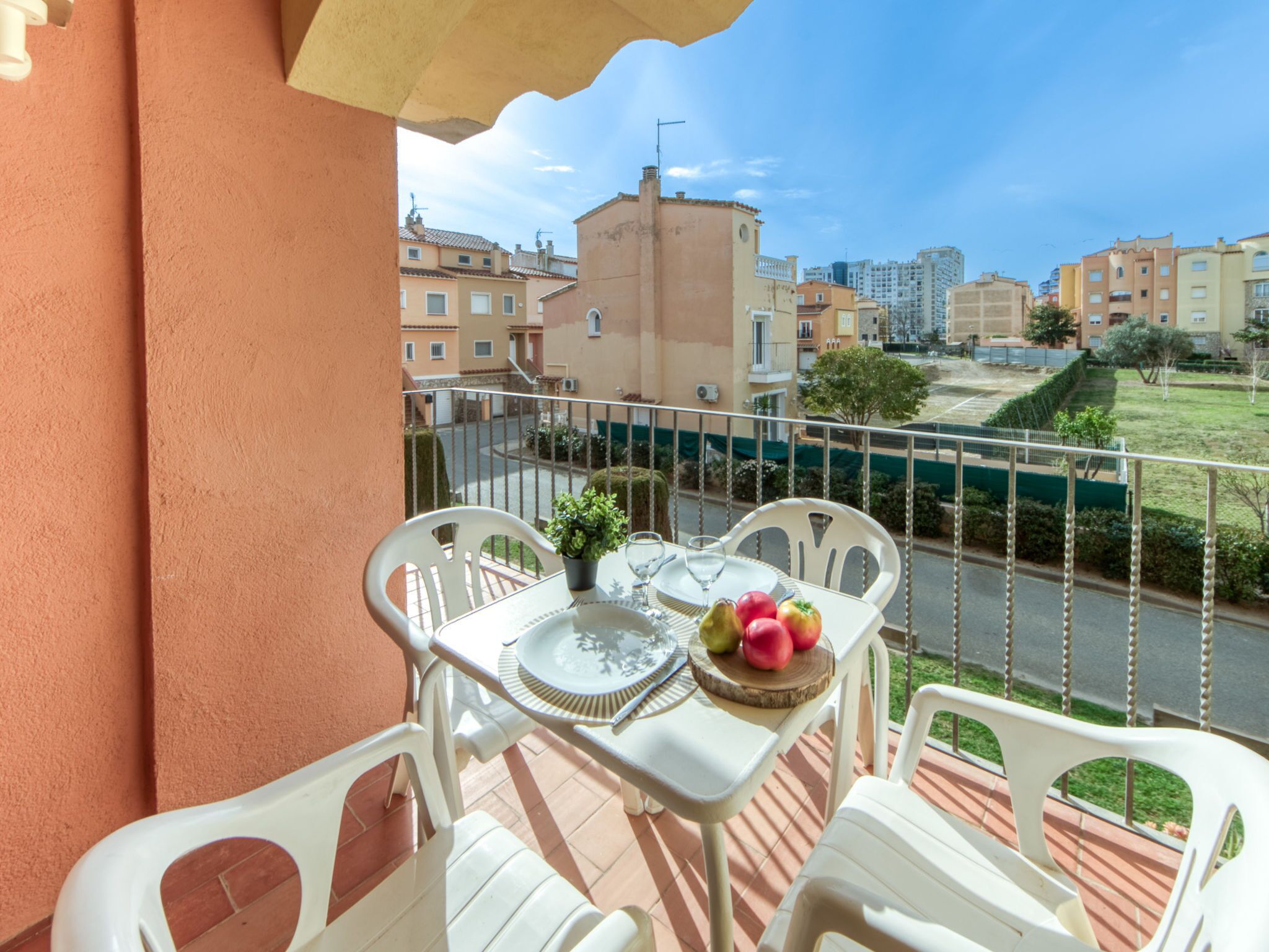 Ein Balkon mit weißen Plastikstühlen, einem Tisch, einem Topfpflanzen und einer Obstschale.