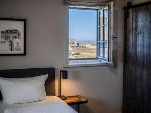 A bedroom with a bed, a bedside table with a lamp, a framed picture on the wall, and a window with a view of houses and a beach.