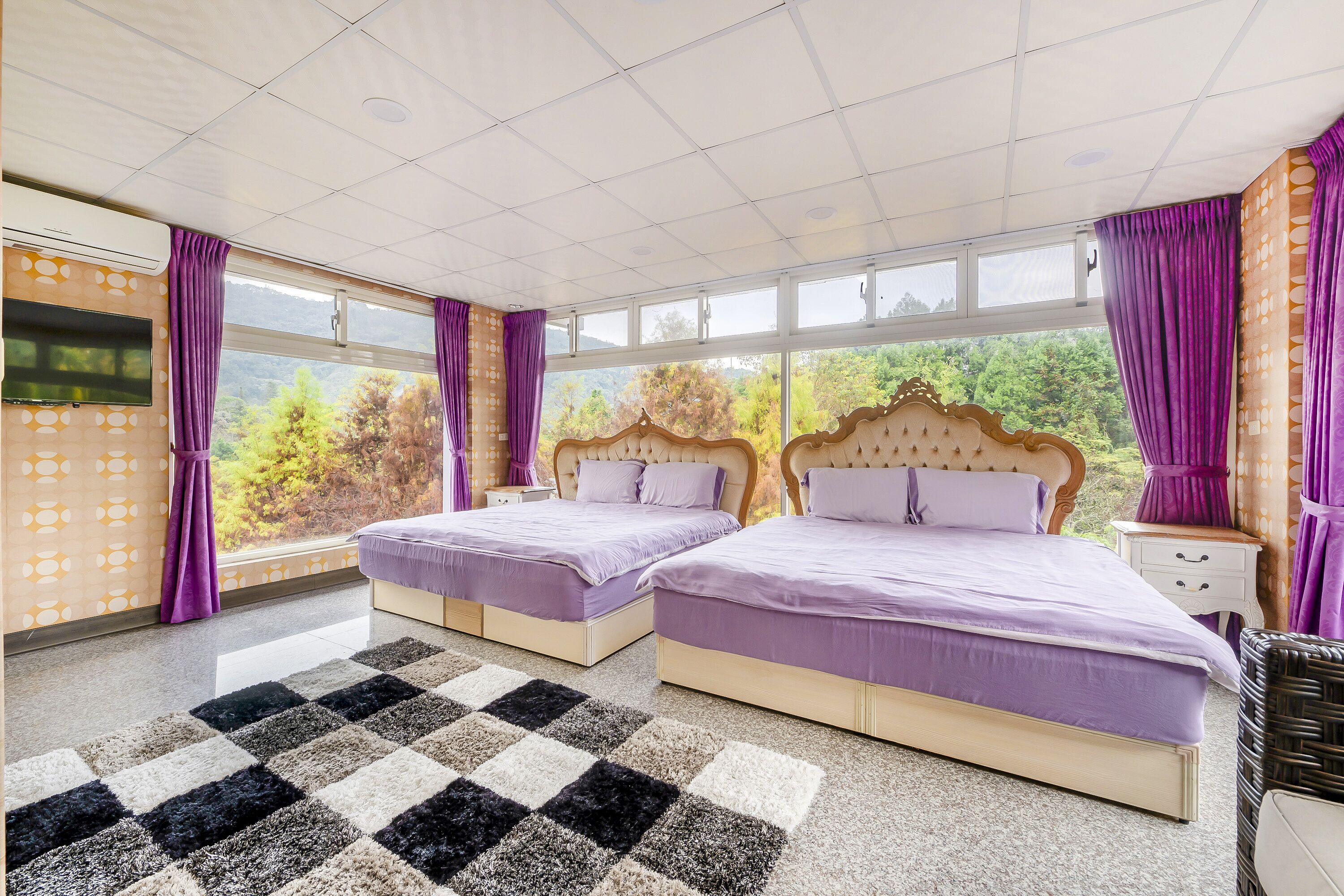 Una habitación de hotel con dos camas, ventanas amplias, un tapete de cuadros y un televisor en la pared.