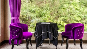 Una habitación con dos sillones de terciopelo morado, una mesa negra cubierta con un mantel blanco y vistas a exuberante vegetación a través de una ventana.