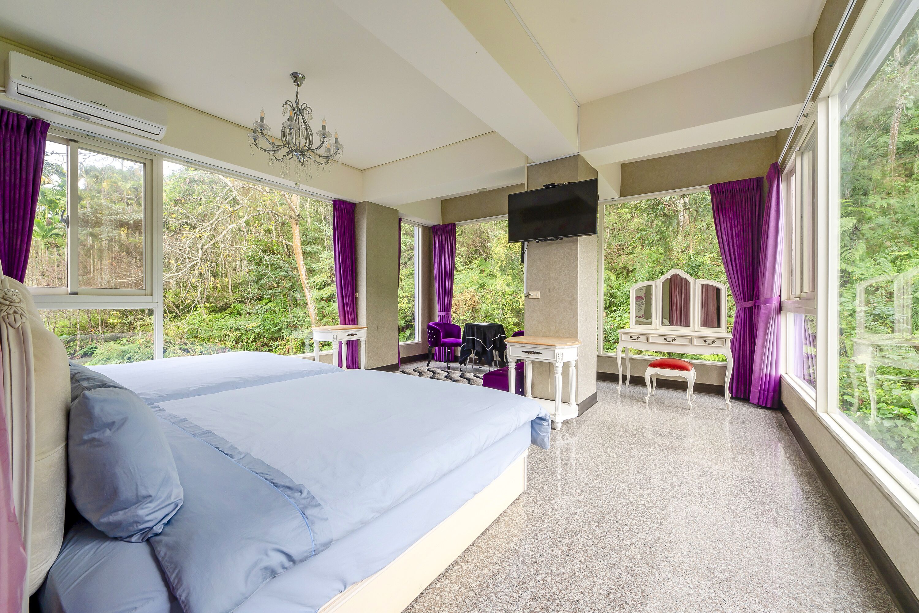 A bedroom with a large bed, a TV, a desk, and a chair, all with large windows offering a view of greenery.