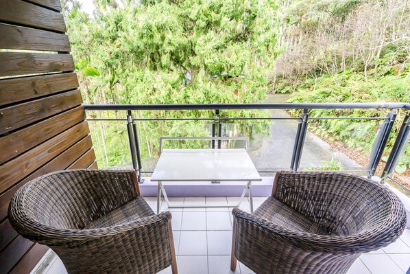 A balcony with wicker chairs and a table, overlooking a lush green area with trees and a path.