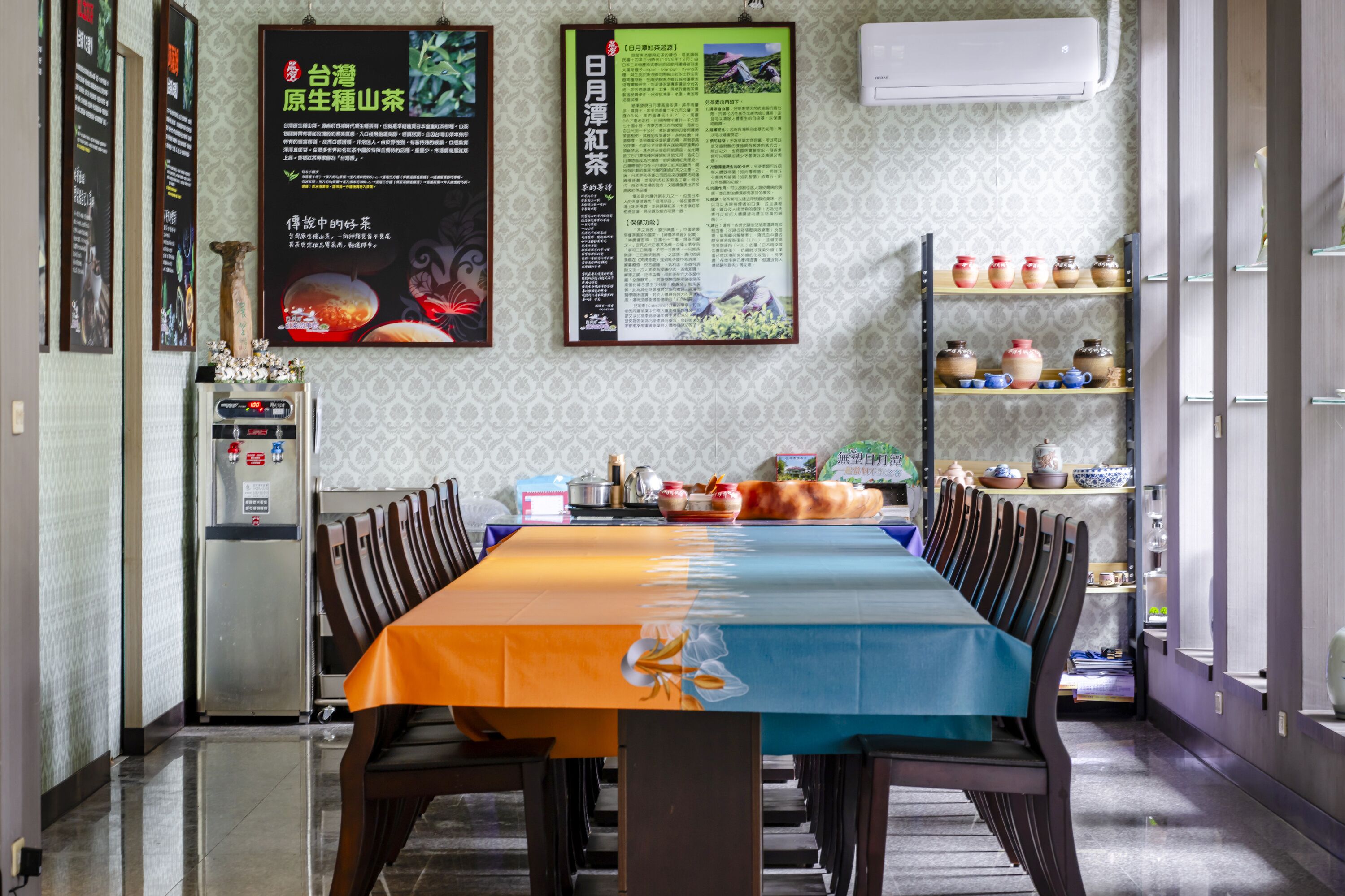 A dining area with a long table, chairs, a refrigerator, and shelves with kitchenware.