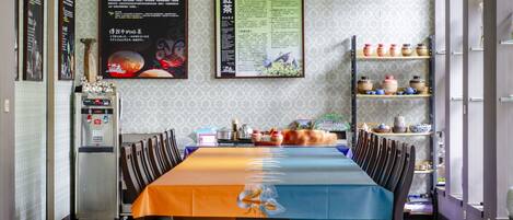 A dining area with a long table, chairs, a refrigerator, and shelves with kitchenware.