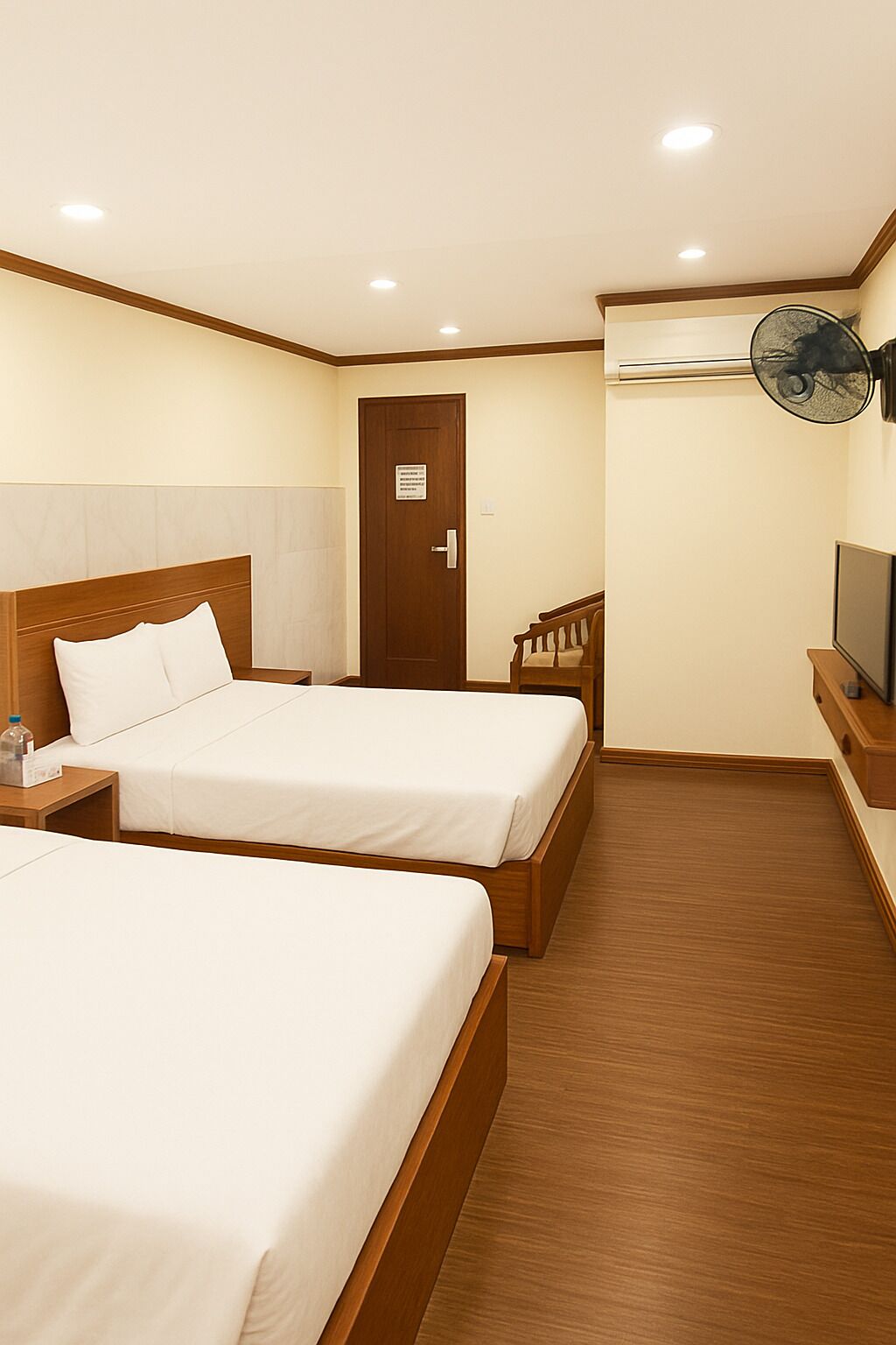 A hotel room with two beds, wooden headboards, a wooden floor, a door, a wall-mounted fan, and a television.