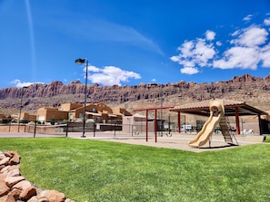 A playground with a slide, surrounded by a fenced area with a building in the background. - Rim Vistas 4a4 | 3 Bed, 2.5 Bath (Moab)