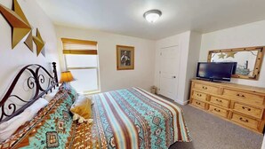Une chambre à coucher comprenant un lit, une commode, un téléviseur et une fenêtre avec des stores.