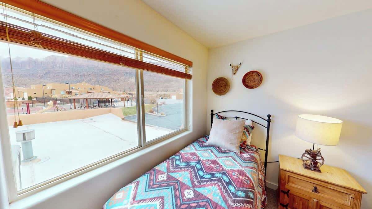 Quarto com cama, mesa de cabeceira com abajur, janela com persianas e vista para um edifício.