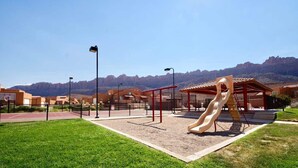 Une aire de jeux avec un toboggan, entourée d’une zone délimitée par une clôture, d’où l’on a une vue dégagée sur les montagnes en arrière-plan.