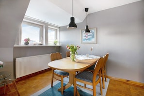 Un coin repas avec une table ronde, quatre chaises, deux lampes suspendues, un vase avec des fleurs, un tableau encadré au mur et une fenêtre donnant sur un paysage.