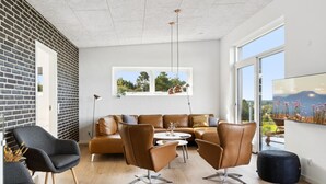 A modern living room with a brown leather sofa, two brown leather chairs, a round coffee table, and a large window with a view of greenery and mountains.