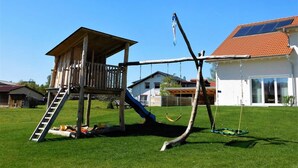Una construcció de fusta per jugar amb tobogan i cadira enfilada, una casa amb teulada vermella i panells solars.