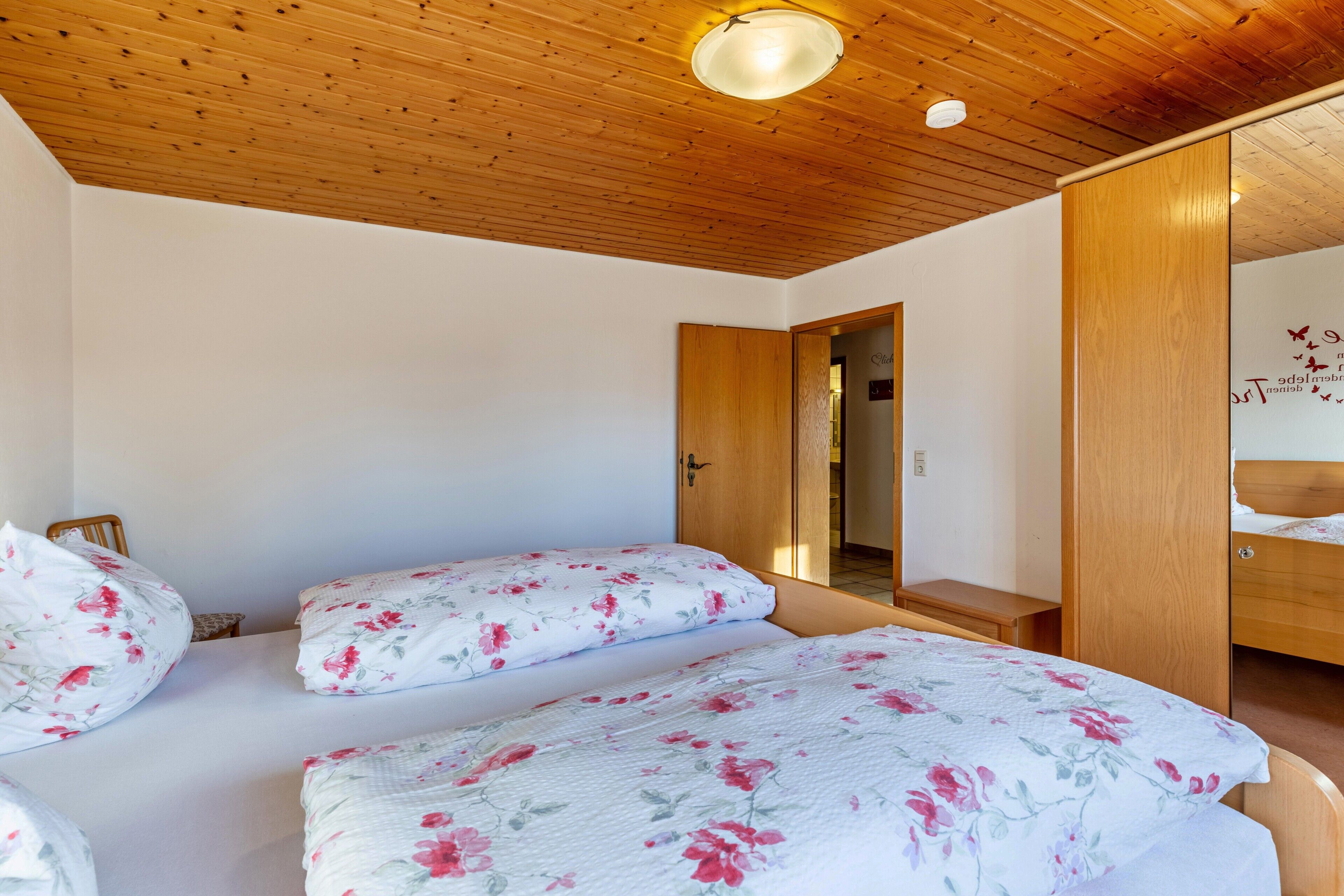 Une chambre avec un plafond en bois, deux lits avec une literie à motifs floraux, une armoire en bois et un miroir.