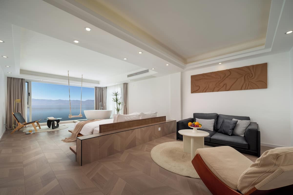Quarto de hotel moderno com uma cama grande, um sofá, uma cadeira balanço e vista para o mar.