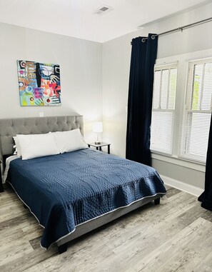 A bedroom with a bed, a nightstand, a lamp, a window with blinds, and a ceiling vent.
