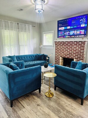 A living room with a blue sofa set, a small round table with a gold base, a brick fireplace, and a mounted TV displaying various channels.