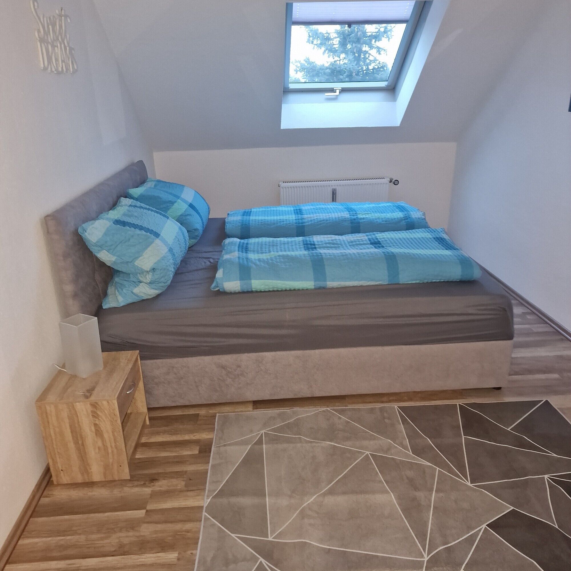 Un dormitorio pequeño y acogedor con tragaluces, una cama con ropa de cama azul y gris, una mesita de noche de madera y una alfombra con estampado geométrico.