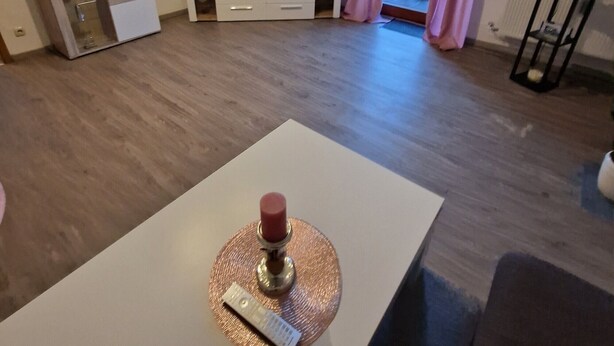Een woonkamer met een flatscreen televisie, een witte salontafel met een kaars, en een balkon met roze gordijnen.