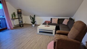 Een woonkamer met een bruine fauteuil, een witte salontafel, een zwarte vloerlamp, een plant in pot en een bank met roze kussens.