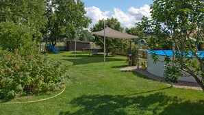 Un patio trasero con césped, un tobogán azul, una zona con sombra y un área de juegos cercada.