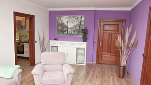 Una sala de estar con un sofá, un comedor con mesa y sillas, una cocina visible a través de una puerta abierta, y una pared morada con una pintura de un paisaje urbano.