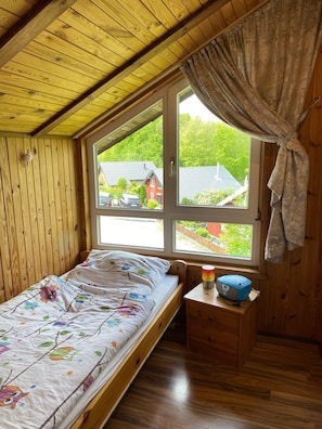 Una habitación tipo cabaña de madera con una cama, una ventana con cortinas, una mesita de noche pequeña con una lámpara y vistas a casas y árboles.
