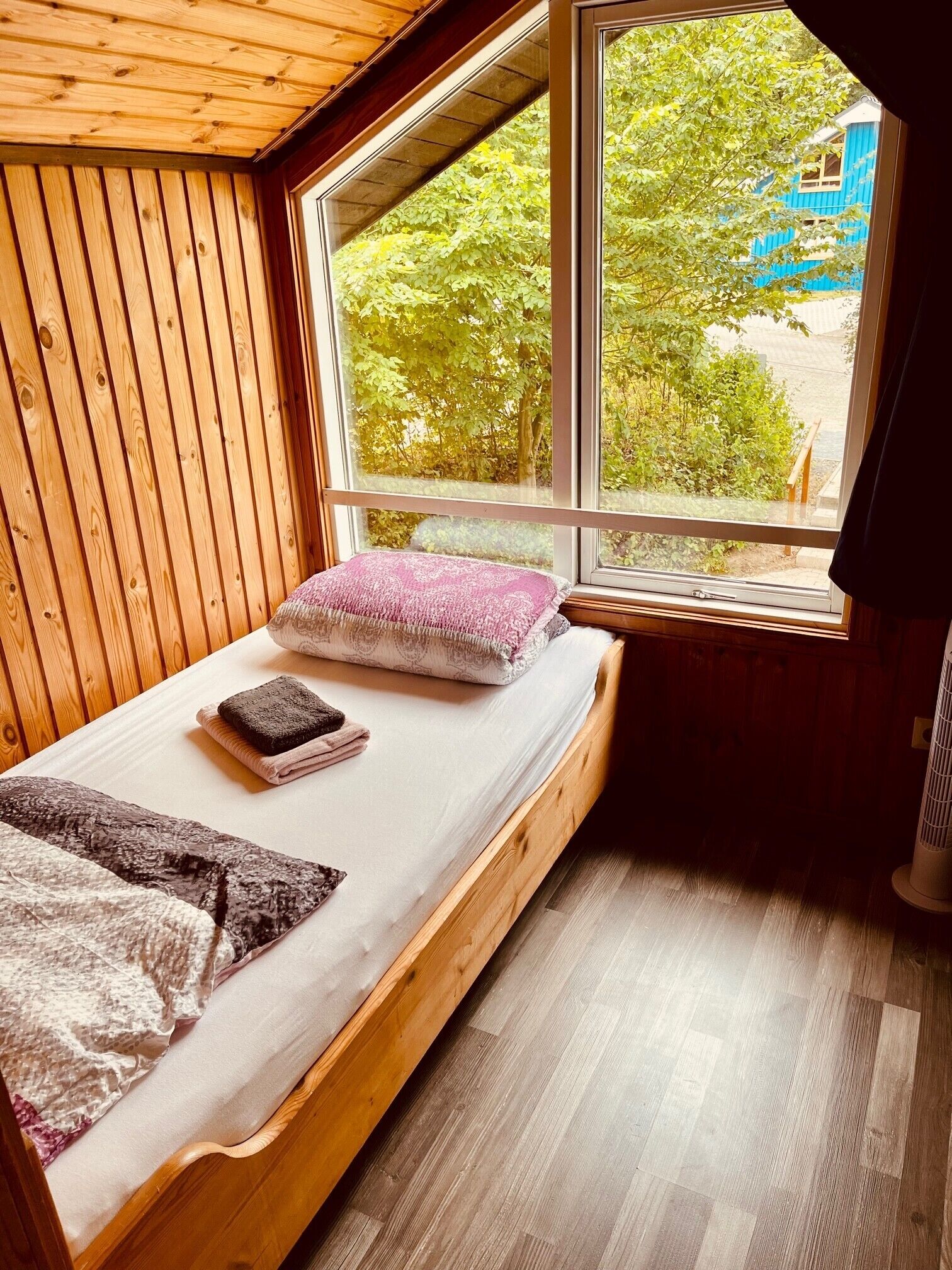 Una pequeña habitación de madera con una cama, un ventanal y un calefactor.