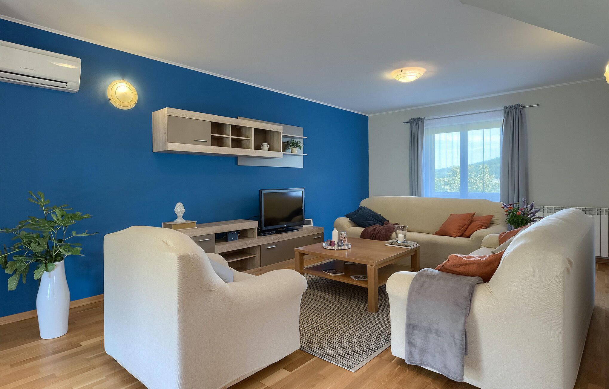 A modern living room with a blue accent wall, a flat-screen TV, a coffee table, two white armchairs, and a beige sofa with orange cushions.