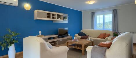 A modern living room with a blue accent wall, a flat-screen TV, a coffee table, two white armchairs, and a beige sofa with orange cushions.