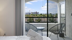 A hotel room with a balcony, a bed with a rolled white towel, two chairs, and a view of the cityscape. - TWO Glendale CozySuites with Pool 2 & 9 (Glendale)