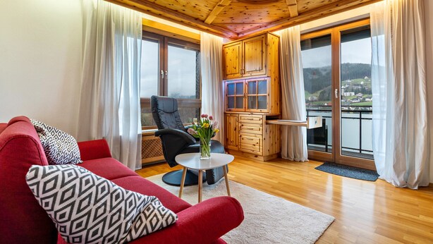 Un salón acogedor con un sofá rojo, una silla negra, una mesita redonda con un jarrón, armarios de madera y ventanas amplias que ofrecen una vista.