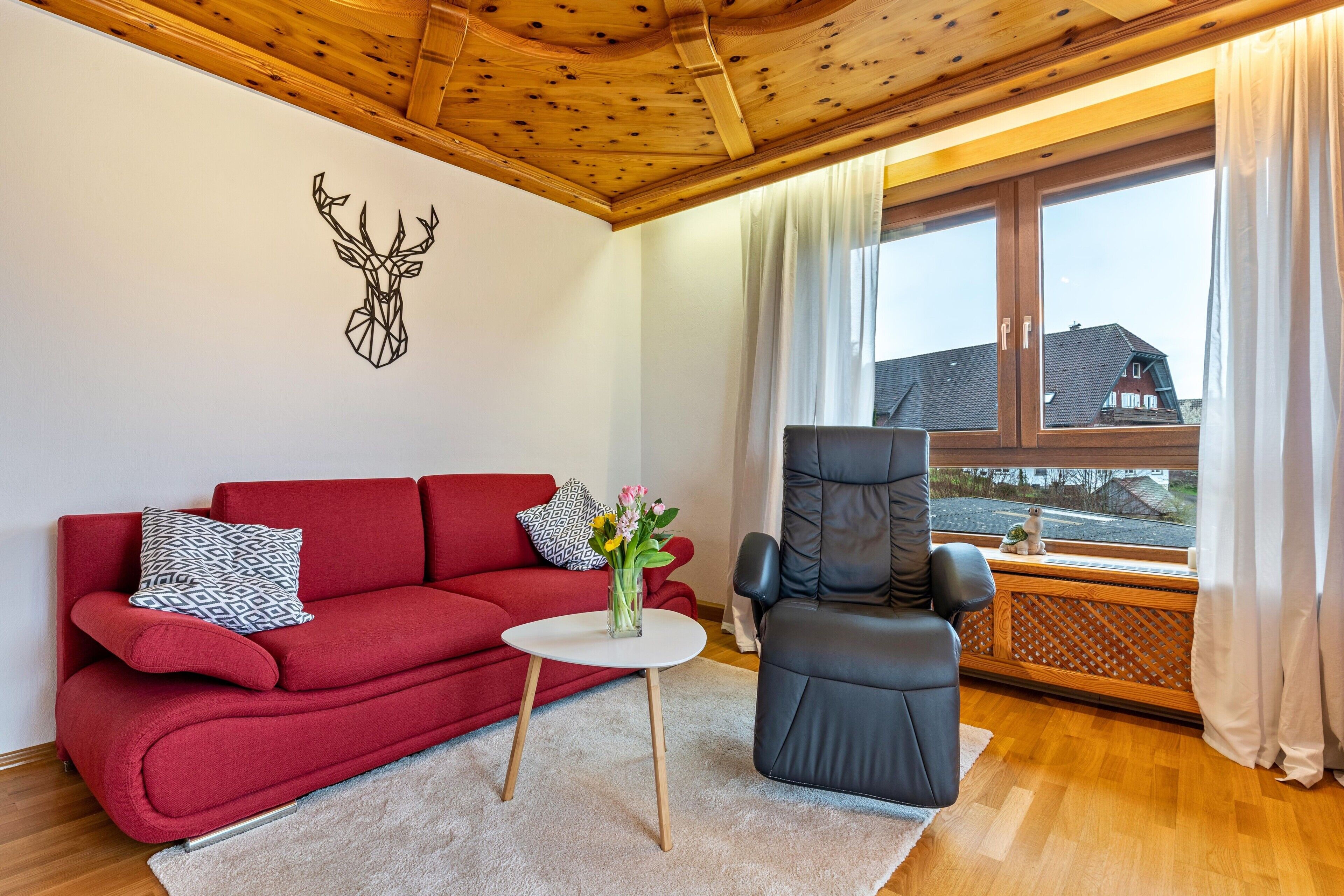 A red sofa, a black chair, a small white table with a vase of flowers, and a wooden ceiling.