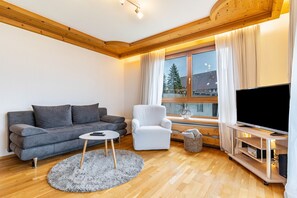 A living room with a grey sofa, a white armchair, a round coffee table, a TV on a wooden stand, and a window with curtains. - Apartment 'Ferienwohnung Schwarzwald' with Mountain View, Shared Terrace and Wi-Fi (Baiersbronn)