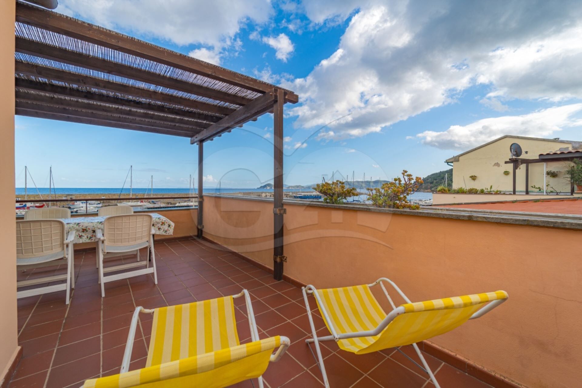 Une terrasse avec deux fauteuils de jardin à rayures jaunes, une table et des chaises. La terrasse donne sur une plage où l’on aperçoit des bateaux et, au loin, un bâtiment.