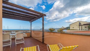 Una terraza con dos sillones reclinables a rayas amarillas, una mesa y sillas. La terraza da a una playa con barcos y un edificio al fondo.