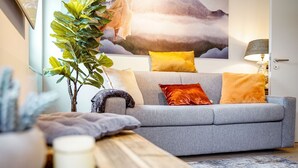 Una sala de estar moderna con un sofá gris, una mesa de centro de madera, una planta en maceta y un cuadro de un paisaje montañoso.