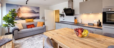 A modern kitchen with a wooden dining table, a fruit bowl, and a grey sofa with orange cushions.