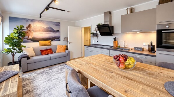 Una cocina moderna con una mesa de comedor de madera, un frutero y un sofá gris con cojines naranja.