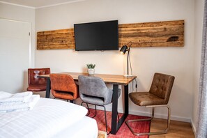 Habitación de hotel con cama, escritorio con dos sillas, televisor montado en una pared de madera y una pequeña planta en maceta.