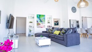 Un salon moderne avec un canapé gris, une table basse blanche et un téléviseur. On y trouve des étagères ornées d’objets décoratifs et un coin repas avec des chaises.