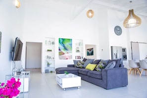 Una sala de estar moderna con un sofá gris, una mesa de centro blanca y un televisor. Hay estanterías con objetos decorativos y un comedor con sillas.