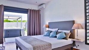 Une chambre à coucher comprenant un lit, des tables de chevet, une fenêtre avec des rideaux, un appareil de climatisation et une vue sur la verdure à l’extérieur.